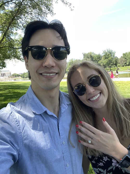 An engaged couple is standing outside on a sunny day, both wearing sunglasses and smiling at the camera. The woman proudly displays an engagement ring on her red-painted left hand, symbolizing their engagement. Green grass, trees, and a large building are visible in the background.